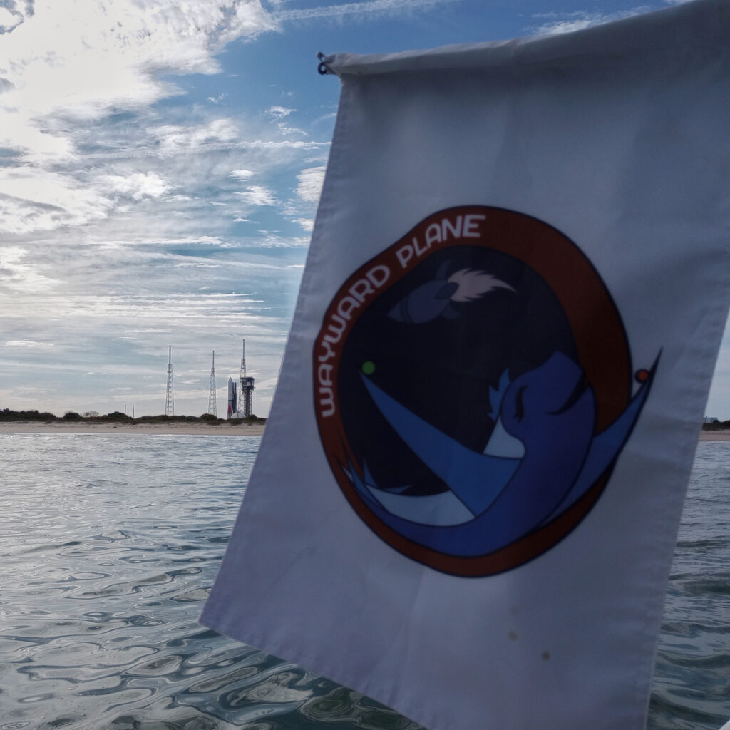 a flag with the personal emblem of WaywardPlane, my deceased roommate, flown off the side of a boat. the Vulcan rocket is seen on the pad in the background on shore.